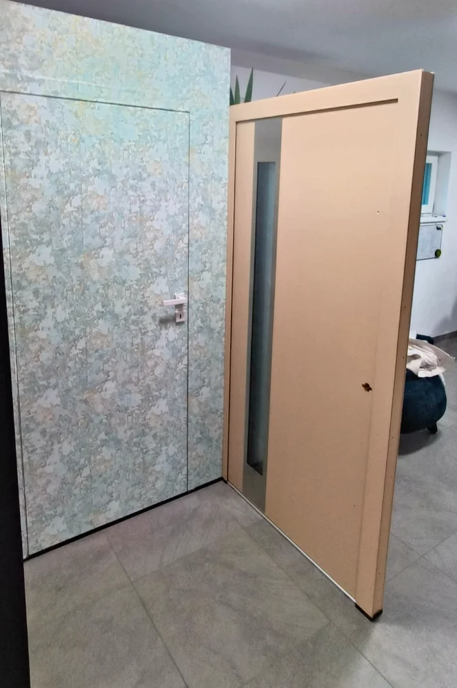 Open peach-pink door with a narrow vertical glass insert, in a hallway with a wallpapered wall